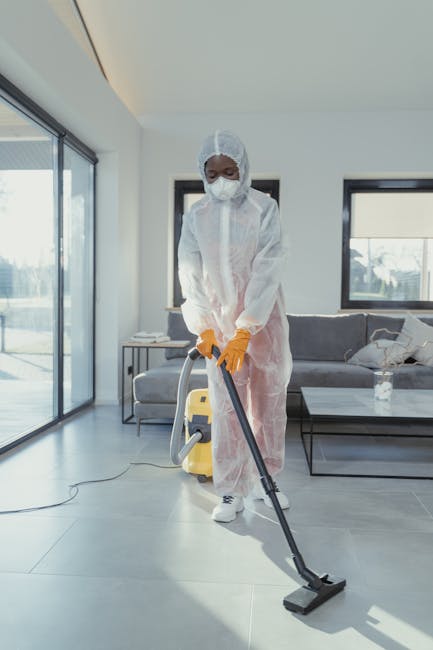 Close-up of a person cleaning a white tiled wall with a blue sponge in their right hand, wearing yellow rubber gloves for surface cleaning and sanitisation. The left hand holds a clear spray bottle fi